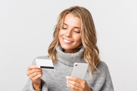 Close-up of blond woman in grey sweater looking at credit card, holding mobile phone while shopping online, standing over white background.