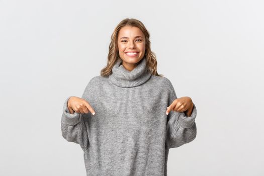 Image of happy woman with short blond hair, wearing grey sweater, pointing fingers down and smiling, standing over white background.
