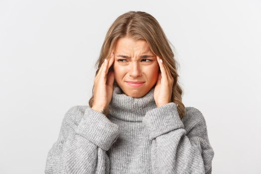 Close-up of grimacing young woman in grey sweater, grimacing and holding hands on head, having painful headache, standing over white background.