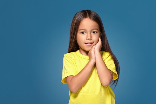 Close up portrait of cute joyful pretty littlegirl with excellent skin and beaming smile, she is admiring her beauty in a mirror, on blue background