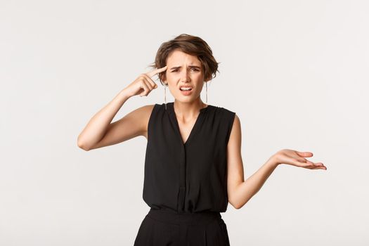 Image of frustrated woman scolding someone, pointing at head and shrugging bothered, standing white background.