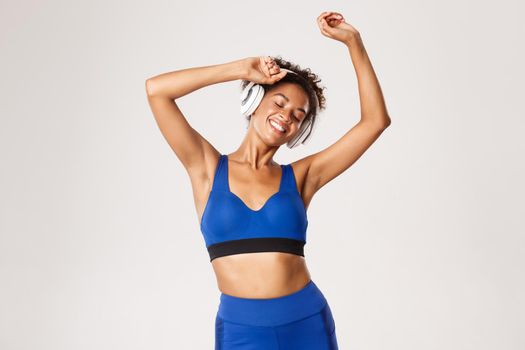 Sport and lifestyle concept. Happy fit african-american woman dancing, smiling with eyes closed, listening music in wireless headphones during workout, white background.