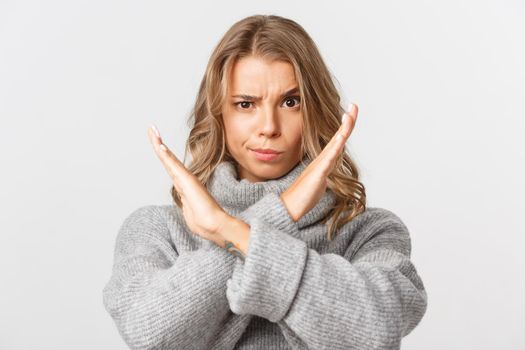 Close-up of serious and confident blond girl in grey sweater, frowning and showing stop gesture, prohibit something, disapprove and forbid action, white background.