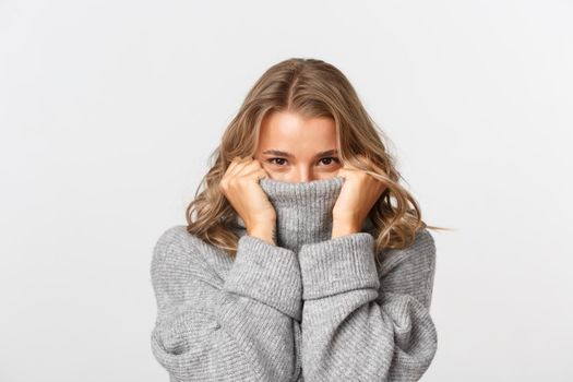Close-up of beautiful blond girl, pulling sweater on face and smiling, standing over white background.