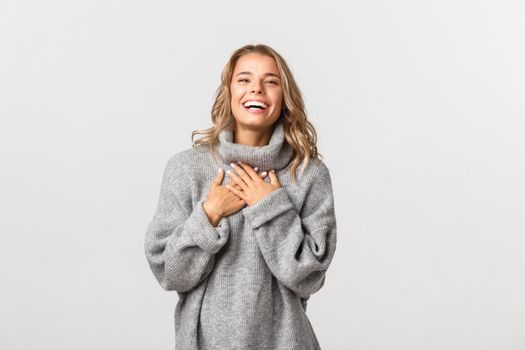 Close-up of happy attractive woman with short blond hair, wearing grey sweater, laughing and looking thankful at camera, standing over white background.