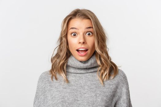 Close-up of attractive blond girl looking surprised, gasping amazed, standing over white background.