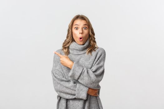 Image of beautiful blond girl in grey sweater pointing finger at upper left corner, showing your logo or banner, looking surprised, standing over white background.