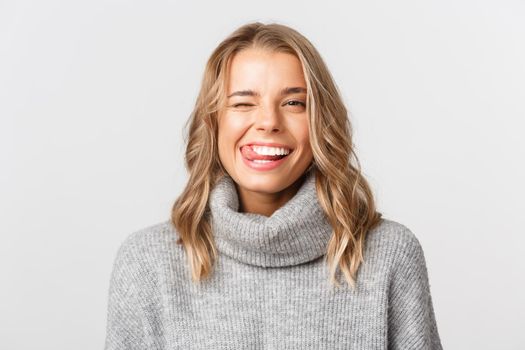 Close-up of happy beautiful blond girl, smiling and showing tongue, winking sassy, standing over white background.