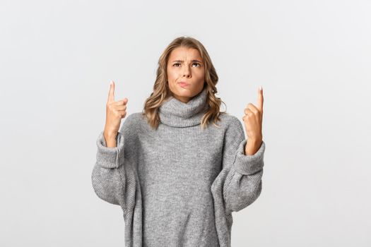 Image of skeptical blond girl in grey sweater, looking indecisive and pointing up, smirking doubtful, standing over white background.