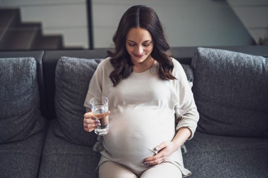 Pregnant woman relaxing at home. She is sitting on bed and drinking water.