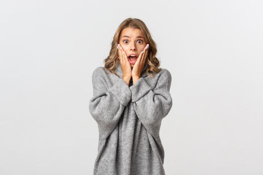 Portrait of scared and worried blond girl in grey sweater reacting to bad accident, screaming and looking concerned, standing over white background.