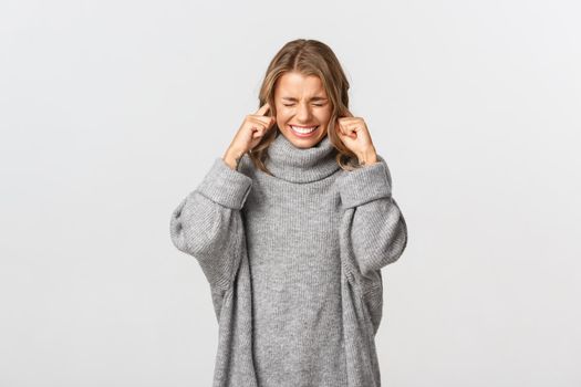 Portrait of annoyed blond girl shut her ears and grimacing from loud noise, standing bothered against white background.