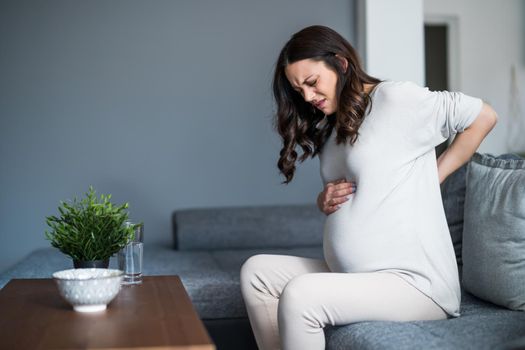 Pregnant woman is having pain in her back. She is sitting on bed at home.