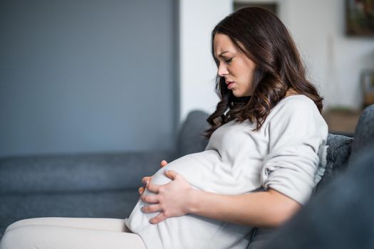 Pregnant woman is having pain in her stomach. She is sitting on bed at home.