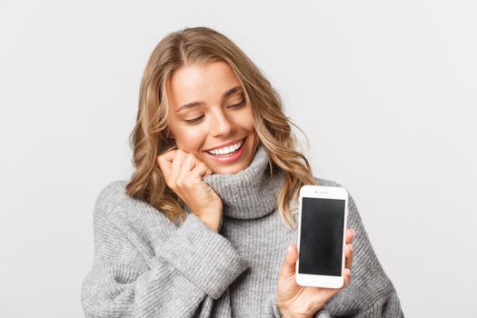 Close-up of lovely blond girl looking happy at mobile phone screen, standing in grey sweater over white background.