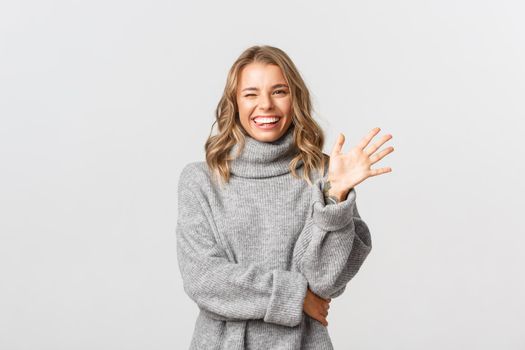 Image of friendly happy woman in grey sweater saying hello, smiling and waving hand to greet person, standing over white background.