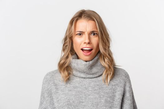 Close-up of attractive blond girl in grey sweater, looking confused at camera, standing over white background.