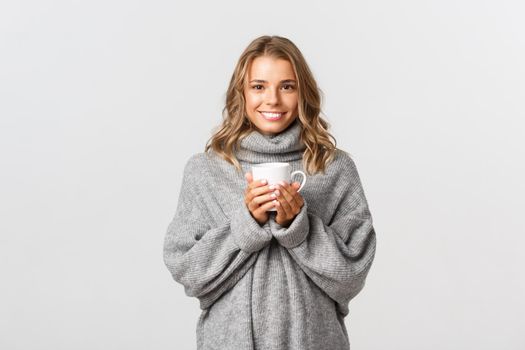 Image of beautiful smiling woman drinking coffee or tea from white mug, standing over white background.