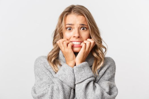 Close-up of scared blond woman in grey sweater, biting fingernails and looking horrified, standing over white background.