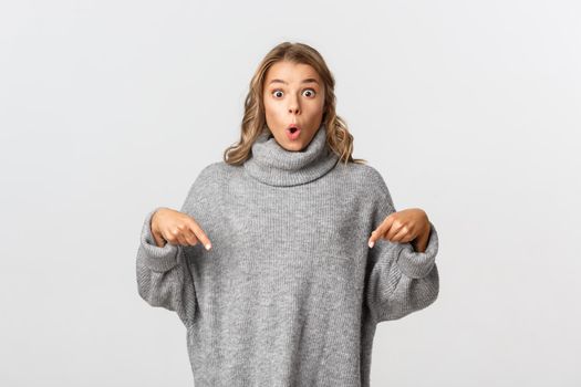 Portrait of surprised girl in grey sweater pointing fingers down, looking impressed with product or brand, white background.