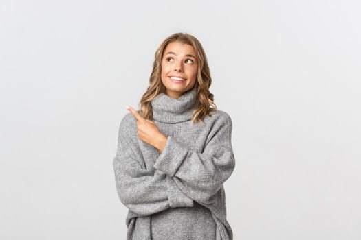 Portrait of attractive blond woman in grey sweater, smiling dreamy and pointing at upper left corner, standing over white background.