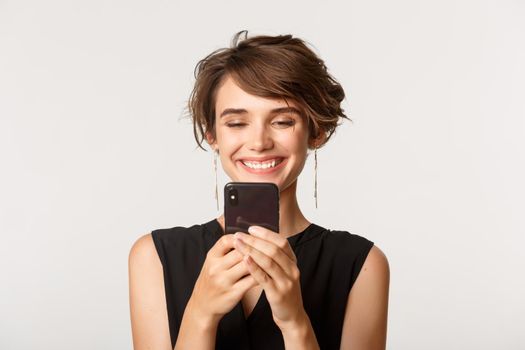 Close-up of satisfied young woman looking at mobile phone with pleased smile, standing over white background.