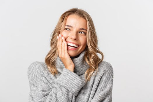 Close-up of attractive happy woman in grey sweater, smiling and looking pleased at upper left corner logo, standing over white background.