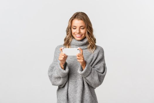 Image of attractive blond girl holding mobile phone horizontally as if watching something or playing games, standing over white background.