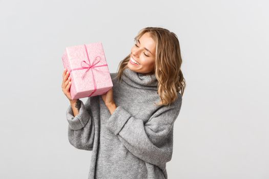 Image of beautiful blond girl in grey sweater, looking happy at birthday gift, receiving present, standing over white background.