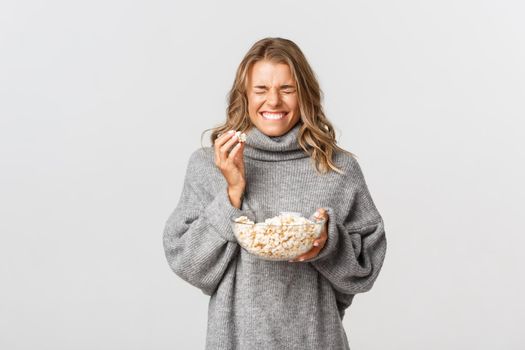 Portrait of happy blond girl, eating popcorn and laughing, watching movie, standing over white background.