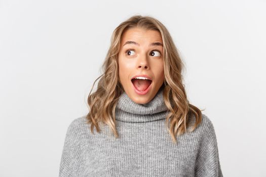 Close-up of amazed blond girl looking at upper left corner, standing surprised over white background.