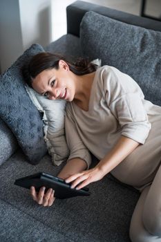 Happy pregnant woman relaxing at home. She is lying on bed in living room and using digital tablet.