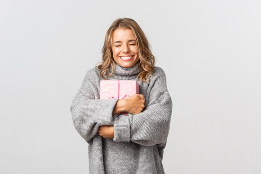 Studio shot of cute smiling girl in grey sweater, hugging her birthday gift, celebrating b-day, standing over white background.