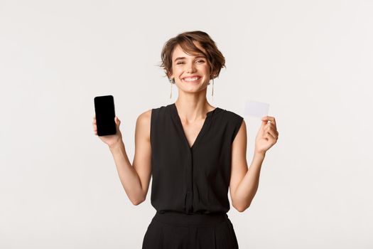 Gorgeous businesswoman showing mobile phone screen and credit card, smiling pleased, standing white background.