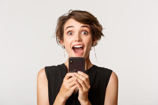 Close-up of excited attractive woman gasping amazed, holding mobile phone, standing over white background.