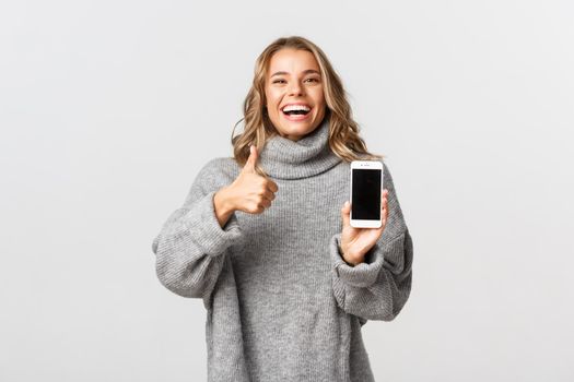 Image of satisfied happy woman in grey sweater, laughing and smiling, showing thumbs-up and mobile phone screen, standing over white background.