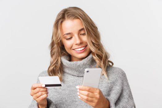 Close-up of attractive blond girl in grey sweater, shopping online with credit card and mobile phone, standing over white background.
