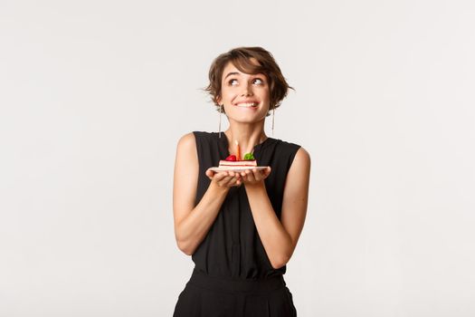 Dreamy cute girl making wish, holding birthday cake and smiling, standing over white background.