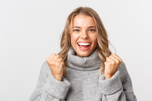 Close-up of hopeful blond girl in grey sweater, clenching fists and looking excited, winning something, standing over white background.