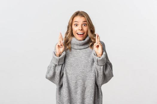 Image of excited and hopeful girl making wish, crossing fingers for good luck, smiling while anticipating results, standing over white background.