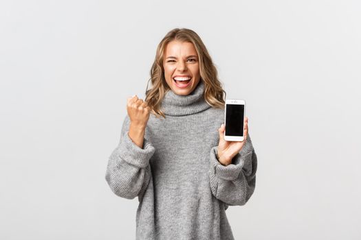 Image of winning happy woman in grey sweater, triumphing over achievement, showing smartphone screen and making fist pump, white background.