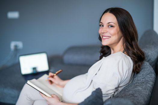 Happy pregnant woman relaxing at home. She is making to do and shopping list.