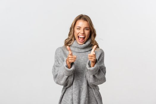 Cheerful smiling female model in casual clothes making choice, need you and pointing at camera, congratulate person, standing over white background.