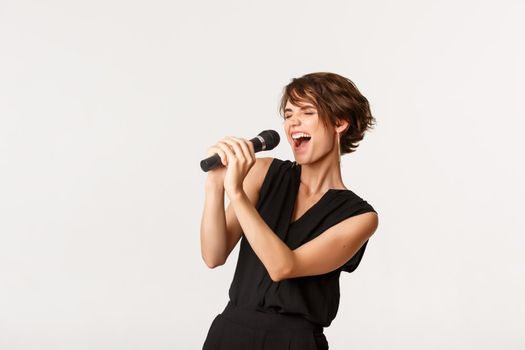 Carefree attractive girl singing in microphone, performing, have fun at karaoke, standing over white background.