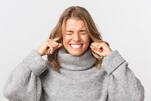 Close-up of annoyed blond girl grimacing, shutting ears and looking up, being irritated by loud noise, standing over white background.