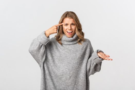 Image of annoyed and frustrated blond girl rolling finger over head, scolding someone for acting stupid, standing over white background.