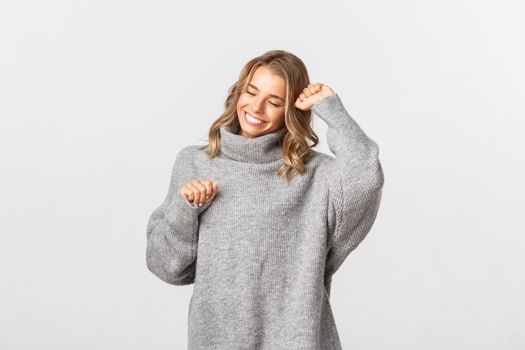 Close-up of attractive blond girl, wearing grey sweater, dancing and smiling happy, standing over white background.