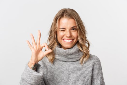 Close-up of confident pretty girl in grey sweater, showing okay sign and smiling, guarantee something, give approval, standing over white background.