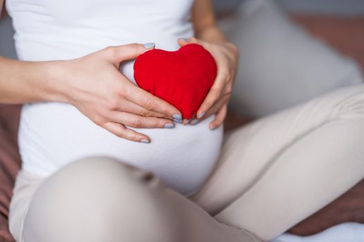 Pregnant woman relaxing at home. She is holding small heart shape pillow on her stomach.