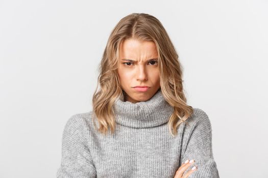 Close-up of angry blond girl in grey sweater, looking from under forehead and sulking, feeling offended, standing over white background.